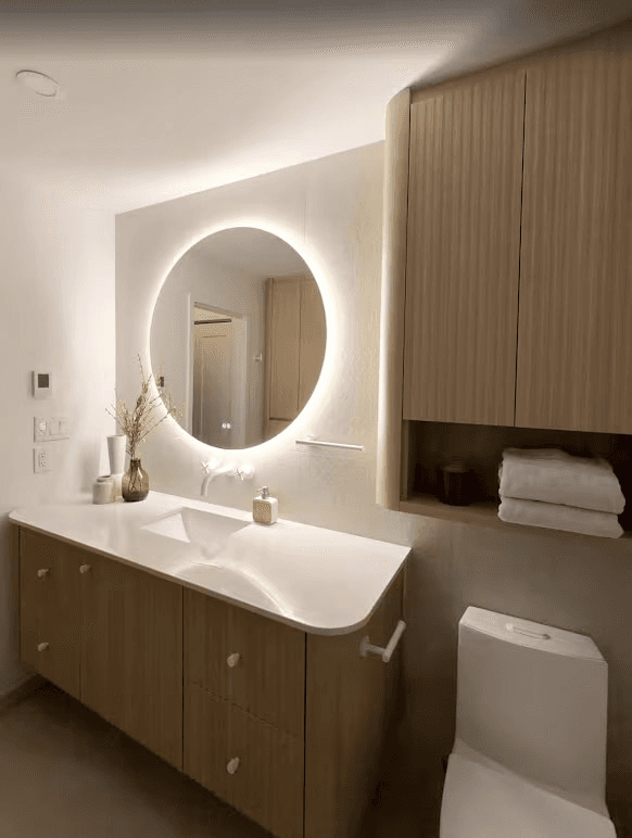 Salle de bain moderne avec vanité suspendue en bois naturel, lavabo intégré au comptoir blanc, miroir rond rétroéclairé DEL, robinetterie murale, armoires de rangement sur mesure en bois rainuré et toilette blanche, au design minimaliste et chaleureux