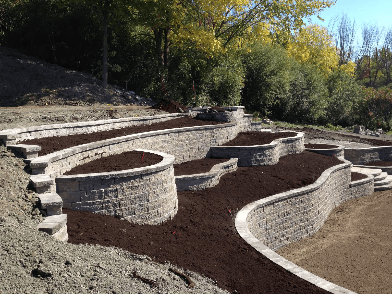 Aménagement paysager en terrasses avec plusieurs murets de soutènement courbés en blocs de béton, escaliers intégrés et zones de plantation sur un terrain en pente