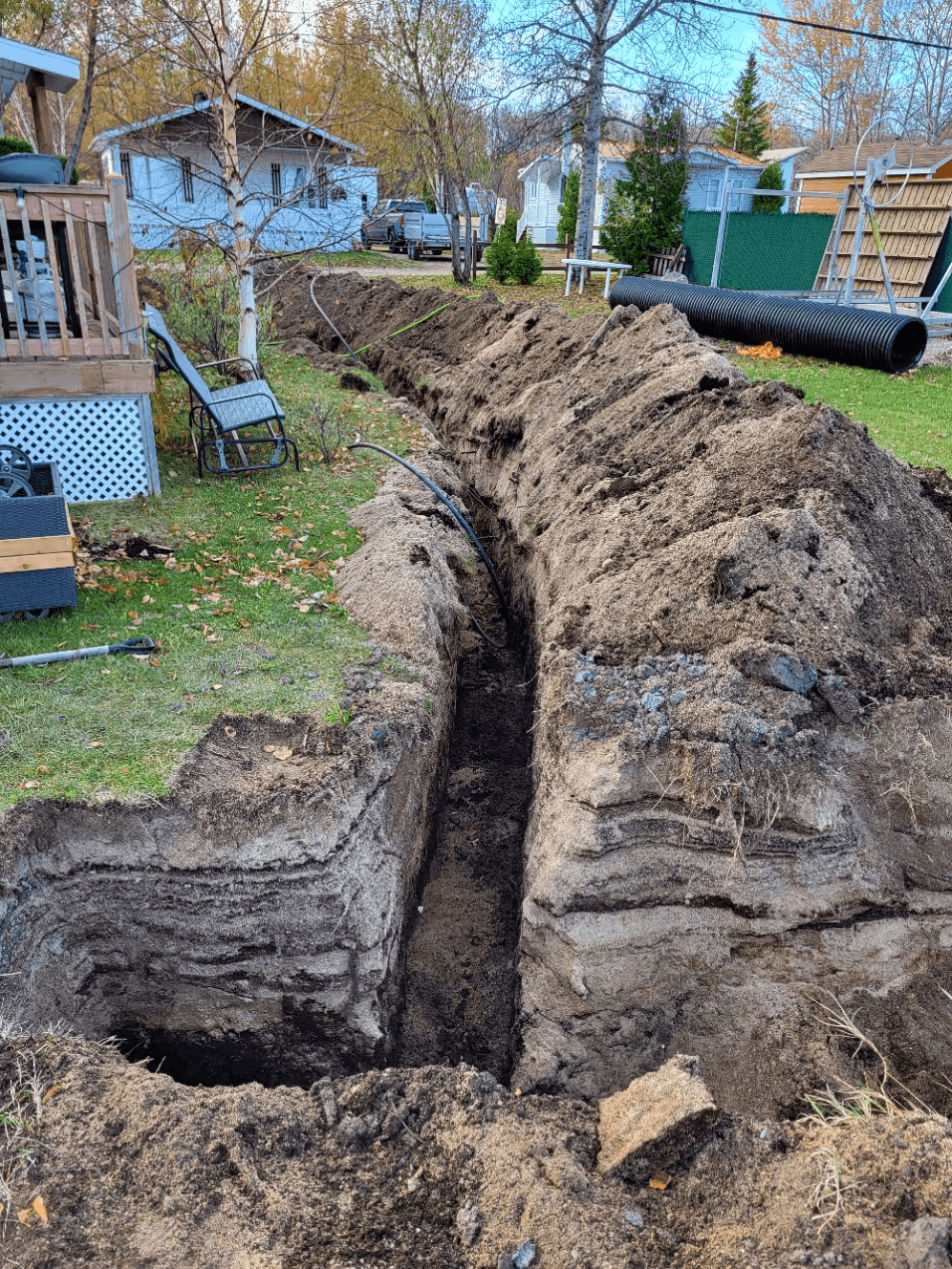 Travaux d’excavation résidentielle dans une cour arrière avec une longue tranchée creusée dans le sol, des conduites noires de drainage, des amas de terre excavée et des tuyaux de drainage annelés, illustrant la préparation d’un système de drainage souterrain sur un terrain résidentiel 