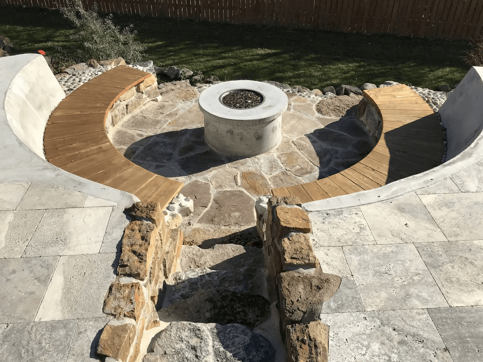 Aménagement extérieur circulaire avec foyer central en béton, bancs courbes en bois, patio en pierre naturelle, murets de maçonnerie et cour gazonnée.