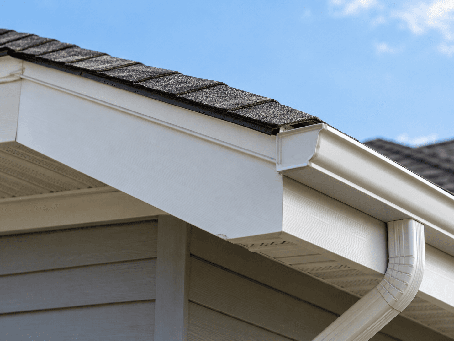 Vue rapprochée d’une toiture résidentielle en bardeaux d’asphalte gris foncé avec gouttière blanche, descente pluviale, fascias blancs et soffites perforés pour la ventilation sous l’avant-toit d’une maison au revêtement extérieur gris