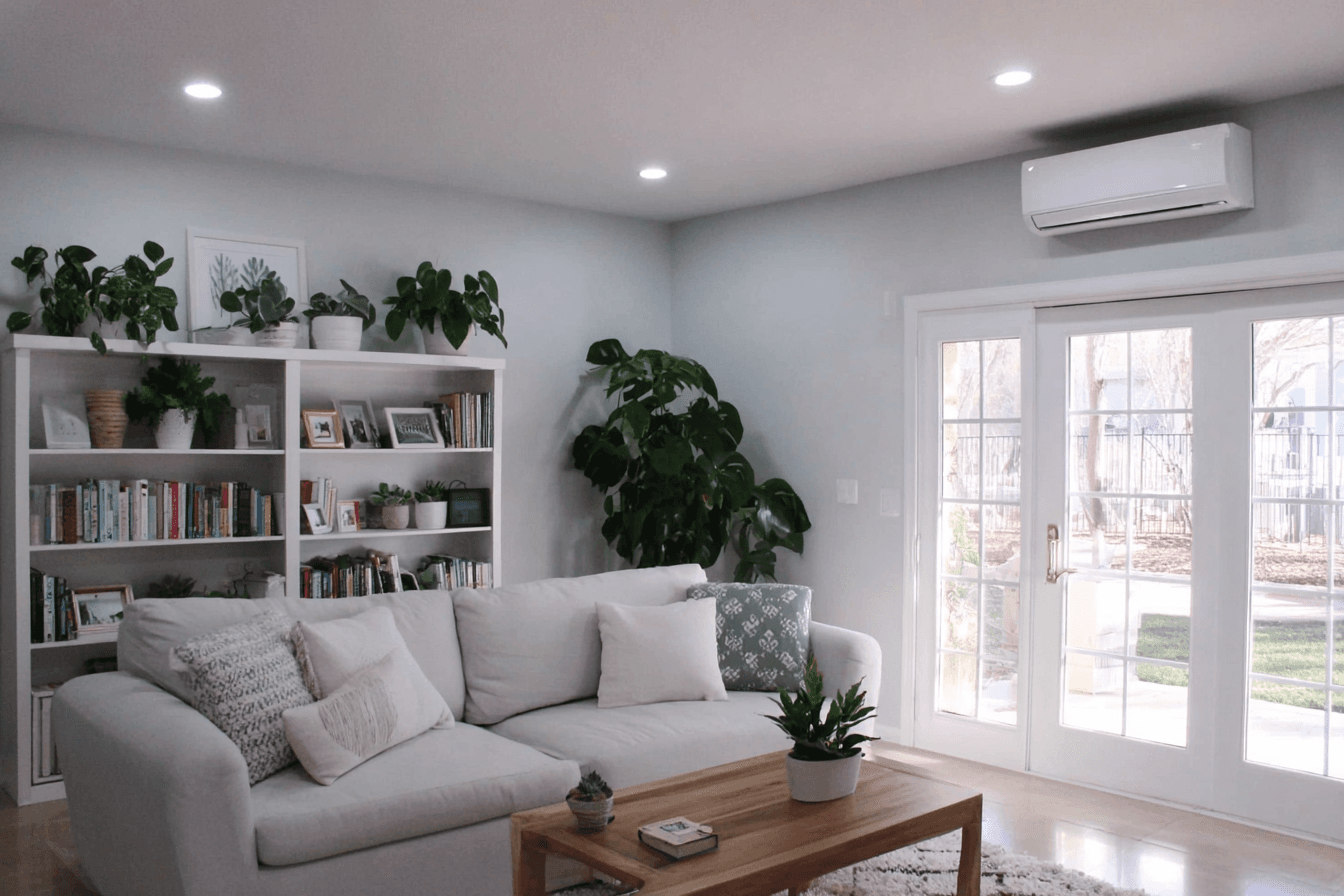 Salon lumineux avec canapé sectionnel blanc, bibliothèque décorée de plantes, portes vitrées et unité murale de thermopompe ou de climatisation