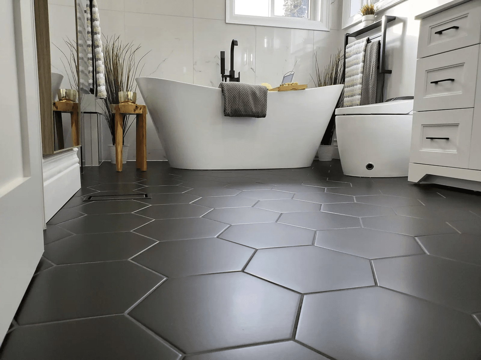 Salle de bain moderne avec baignoire autoportante blanche, robinetterie noire mate, plancher en céramique hexagonale gris anthracite, murs en carreaux effet marbre blanc, vanité blanche avec poignées noires, toilette contemporaine et fenêtre apportant une lumière naturelle