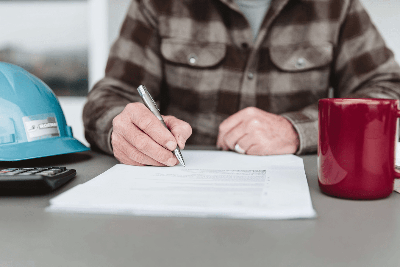 Professionnel de la construction remplissant ou signant un document à une table, avec casque de chantier, calculatrice et tasse à proximité