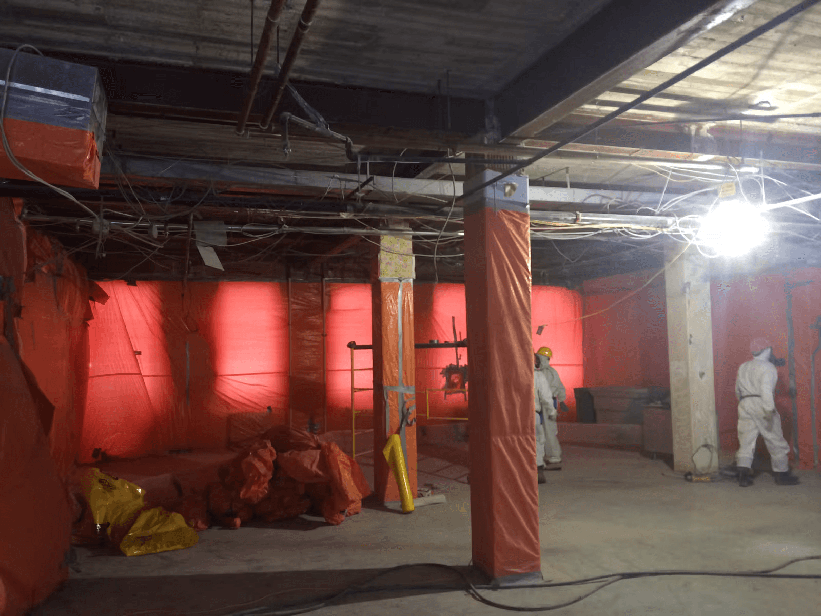 Chantier de décontamination intérieure dans un bâtiment commercial avec deux travailleurs en combinaisons de protection blanches, murs et colonnes recouverts de bâches plastiques orange, câbles apparents au plafond et sacs de déchets, illustrant des travaux spécialisés de confinement et de retrait de matériaux potentiellement dangereux