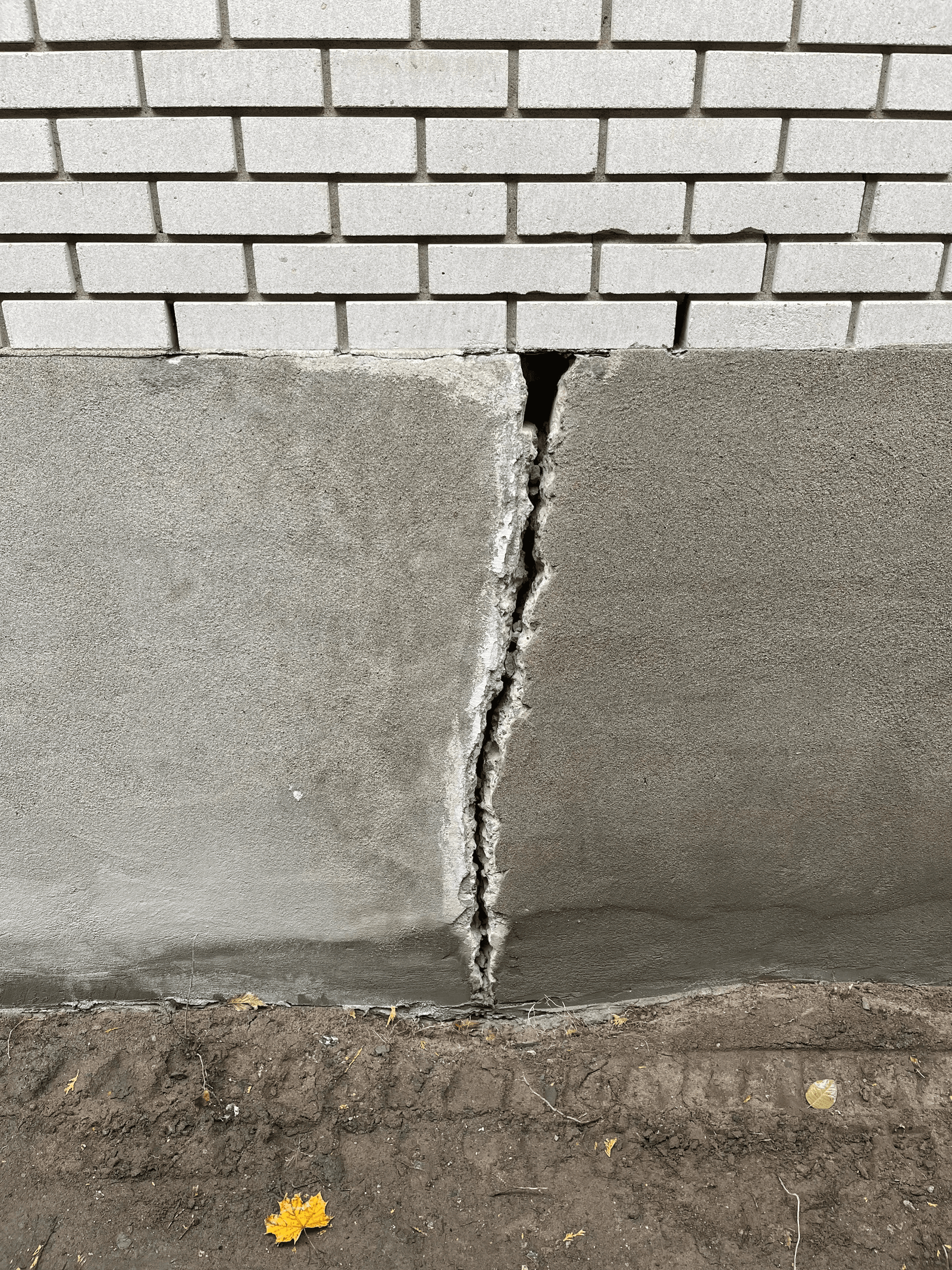 Fissure verticale importante dans un mur de fondation extérieur sous un parement de brique, avec béton endommagé nécessitant une réparation structurale et une imperméabilisation / Large vertical crack in an exterior foundation wall beneath brick cladding, with damaged concrete requiring structural repair and waterproofing