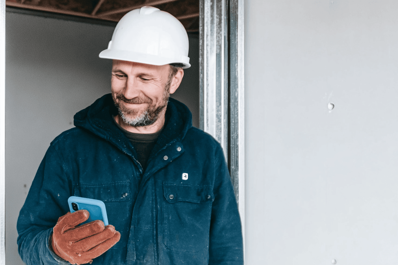  Entrepreneur en construction portant un casque blanc et des gants de travail, consultant son téléphone à l’intérieur d’une maison en chantier
