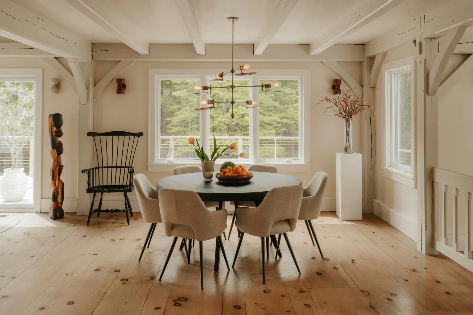 Salle à manger lumineuse au style chaleureux avec table ronde, chaises rembourrées, poutres apparentes, plancher de bois et grandes fenêtres donnant sur la nature
