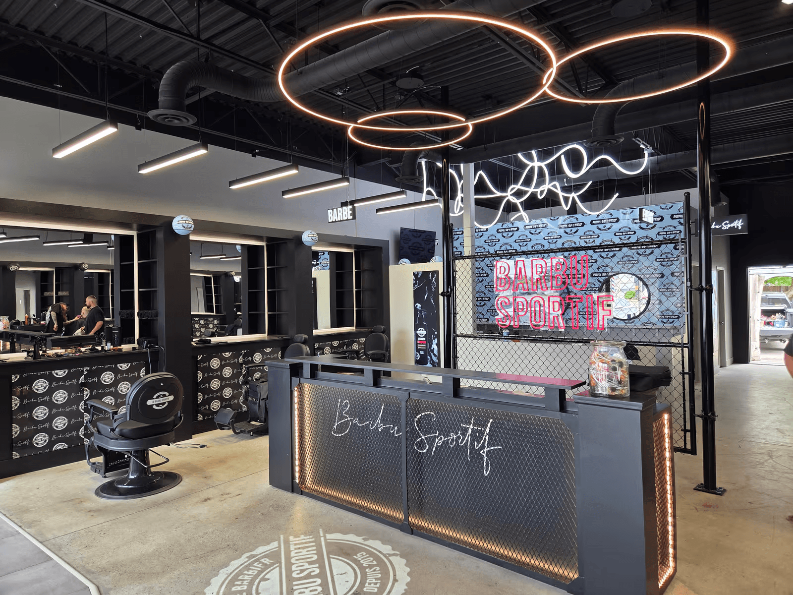 Intérieur moderne d’un salon de barbier avec postes de coiffure, fauteuils, comptoir d’accueil, éclairage suspendu circulaire et décor industriel