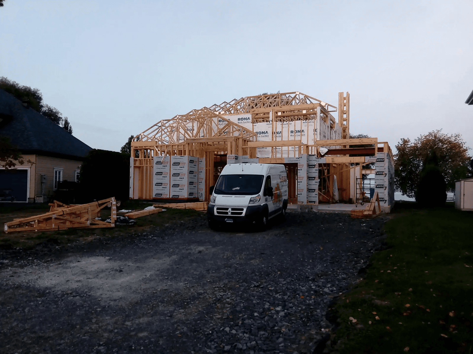 Maison en cours de construction avec charpente en bois, revêtement pare-intempéries RONA et véhicule de chantier stationné sur l’allée en gravier.