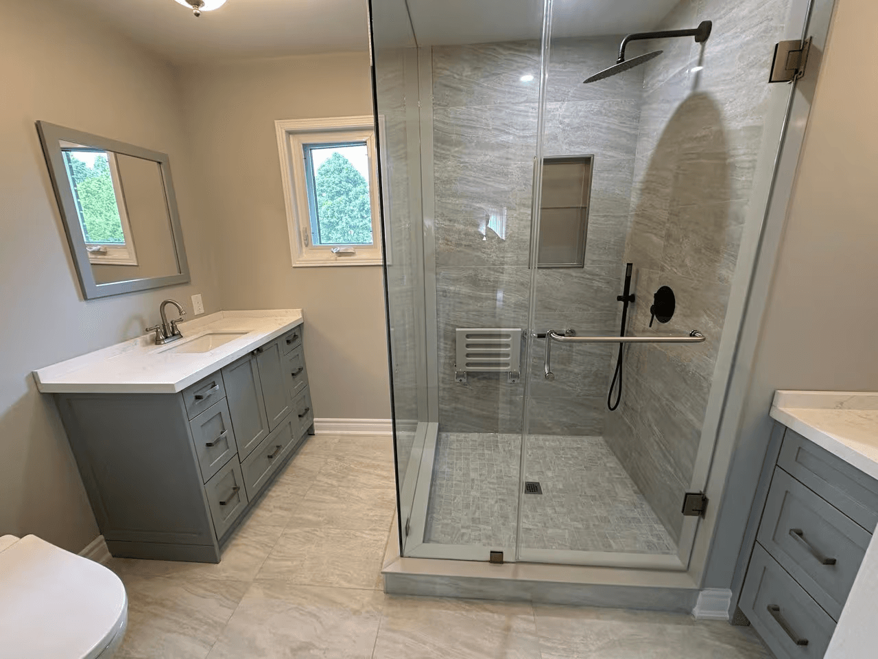 Salle de bain rénovée avec grande douche vitrée en céramique grise, douche pluie noire, vanités grises et comptoirs blancs