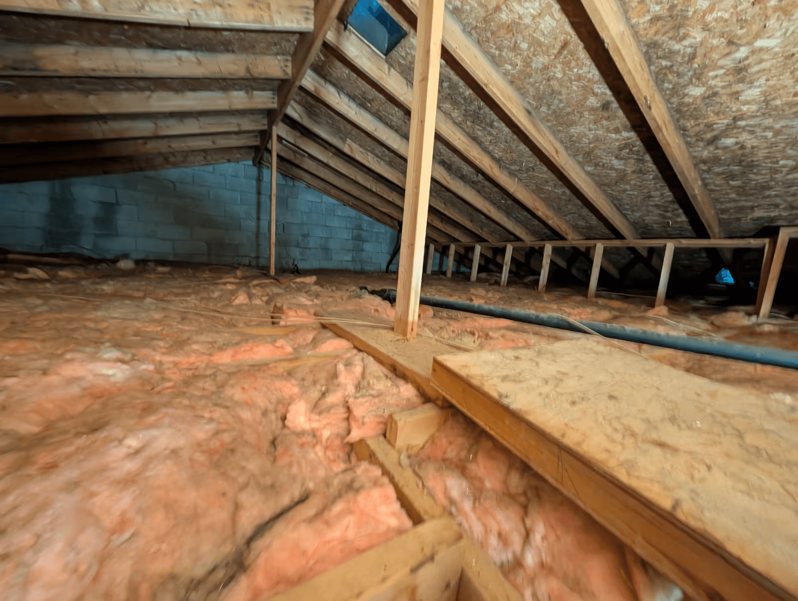 Entretoit résidentiel avec charpente en bois apparente, chevrons et fermes de toiture, panneaux de contreventement en OSB, isolation en laine de fibre de verre rose soufflée ou déposée entre les solives, mur de blocs de béton, conduits de ventilation et plateforme d’accès en bois