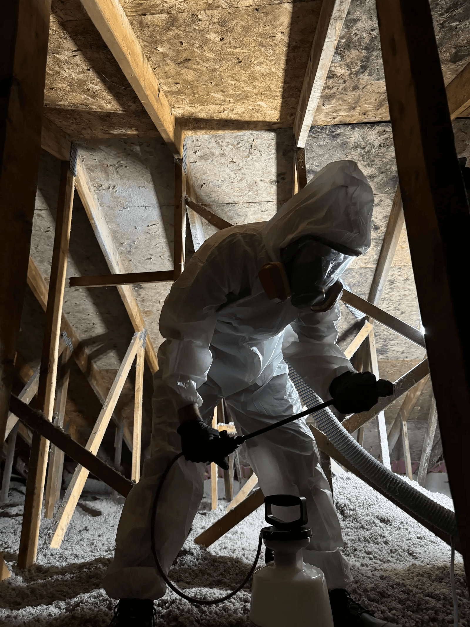 Technicien en combinaison de protection blanche, masque respiratoire à filtres et gants noirs utilisant un pulvérisateur manuel dans un entretoit résidentiel avec charpente en bois et isolation soufflée, illustrant une intervention spécialisée de traitement et de décontamination des combles