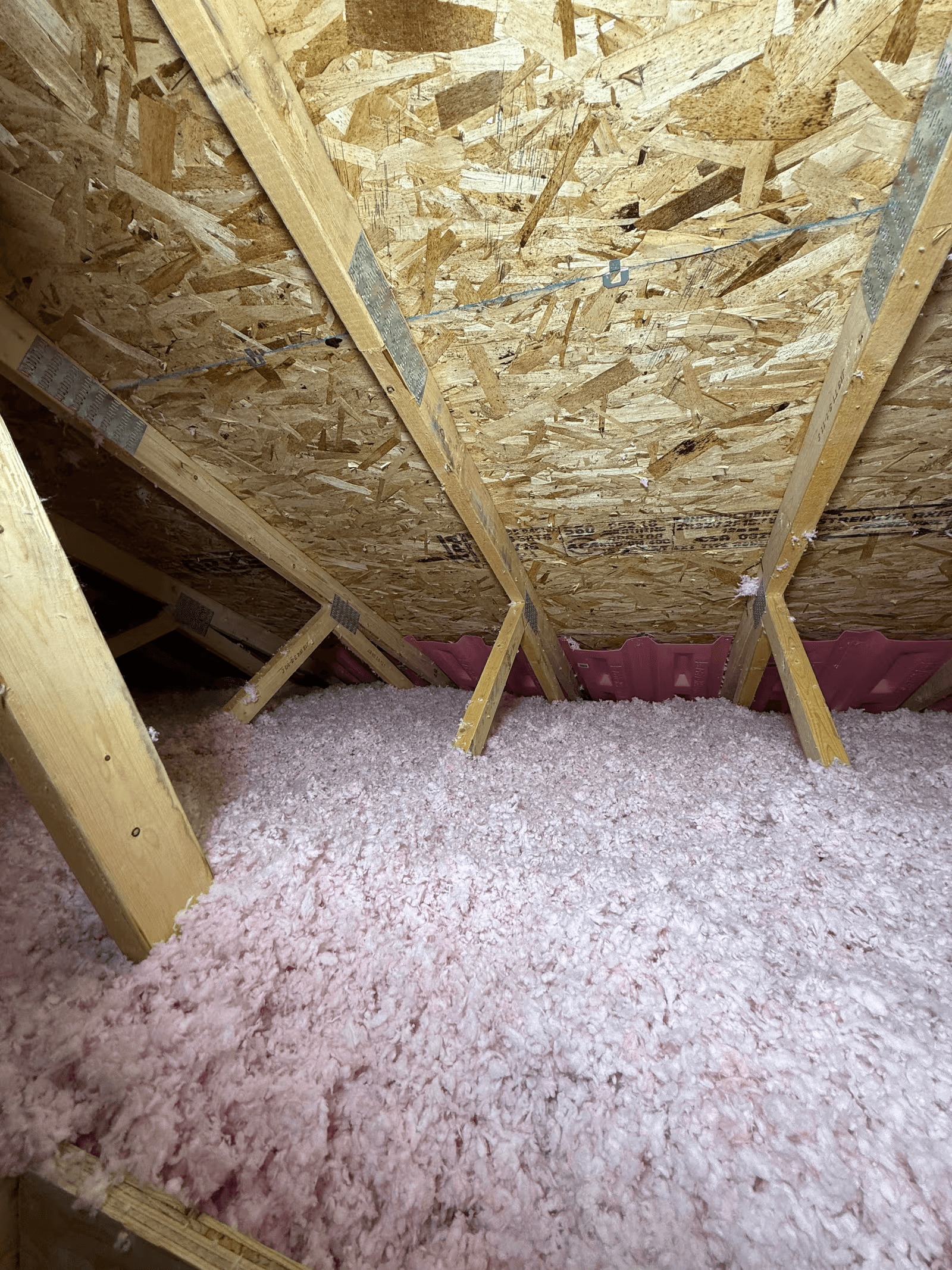 Entretoit résidentiel isolé avec une épaisse couche de laine isolante soufflée rose et blanche, charpente de toiture en bois avec fermes triangulées, panneaux OSB apparents, connecteurs métalliques et déflecteurs de ventilation roses installés près des avant-toits
