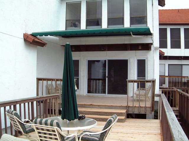 Auvent rétractable en tissu vert installé au-dessus de la porte-fenêtre d'une terrasse en bois avec mobilier de patio.