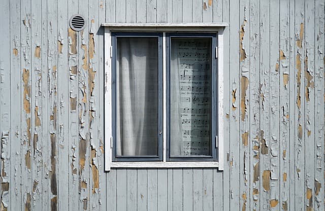 Fenêtre en bois sur un mur de façade à la peinture grise écaillée nécessitant des travaux de rénovation.